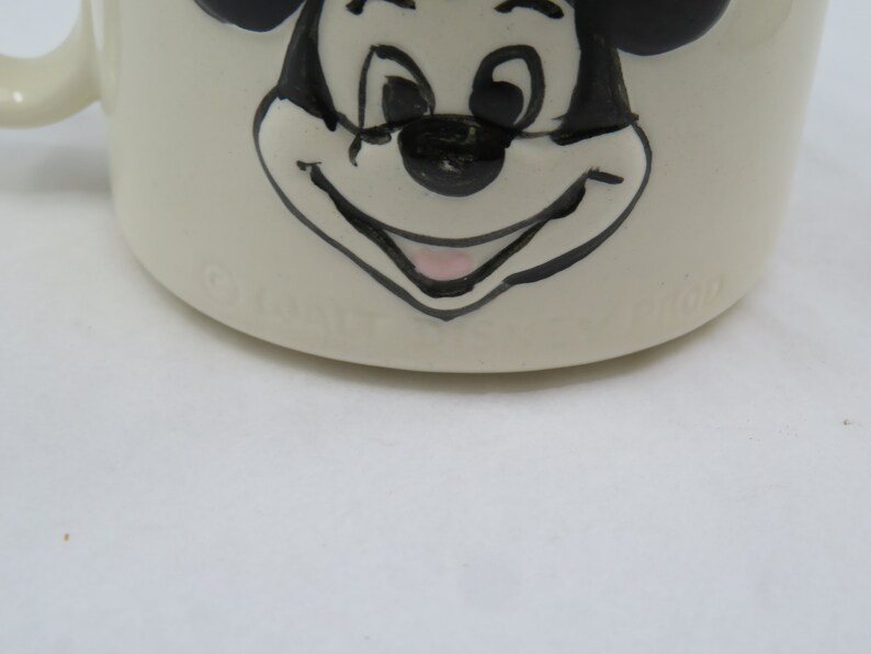 May include: White ceramic mug with a black and white illustration of Mickey Mouse's face. The mug has the word "Disney" printed on the bottom.