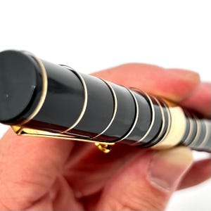 Puede incluir: Primer plano de un bol&iacute;grafo negro y dorado. El bol&iacute;grafo tiene un cuerpo negro con detalles dorados y un clip dorado. El bol&iacute;grafo se sostiene en una mano sobre un fondo blanco.
