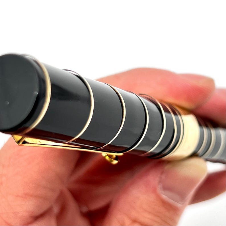 Puede incluir: Primer plano de un bol&iacute;grafo negro y dorado. El bol&iacute;grafo tiene un cuerpo negro con detalles dorados y un clip dorado. El bol&iacute;grafo se sostiene en una mano sobre un fondo blanco.