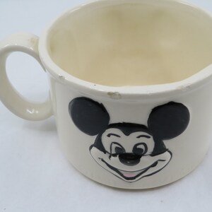 May include: White ceramic mug with a black and white image of Mickey Mouse. Mickey Mouse is smiling with his tongue sticking out.