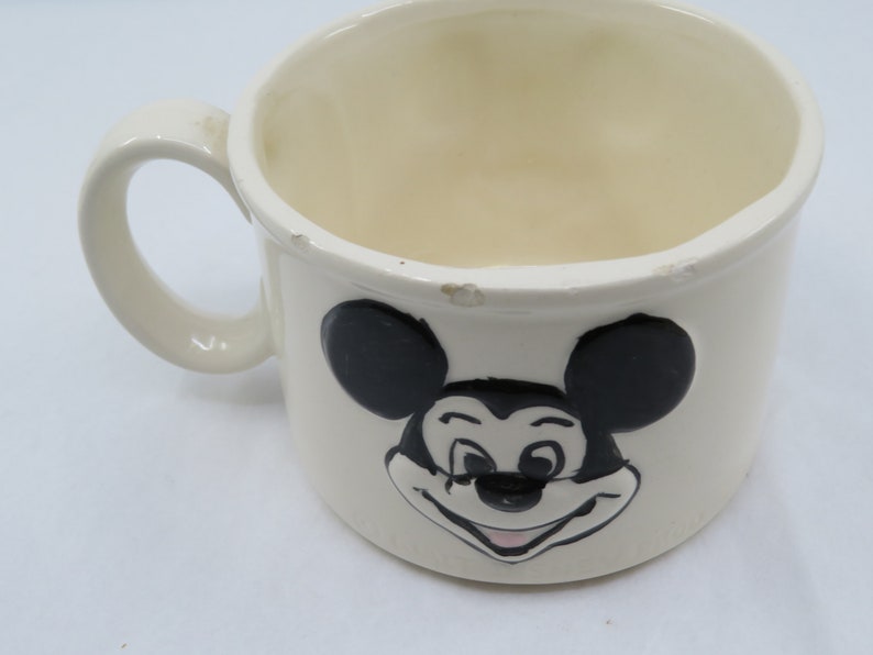 May include: White ceramic mug with a black and white image of Mickey Mouse. Mickey Mouse is smiling with his tongue sticking out.