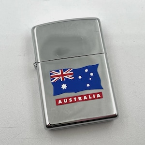 May include: A silver-colored lighter with the Australian flag design. The flag is blue with the Union Jack in the upper left corner, a constellation of white stars, and the word "AUSTRALIA" in red.
