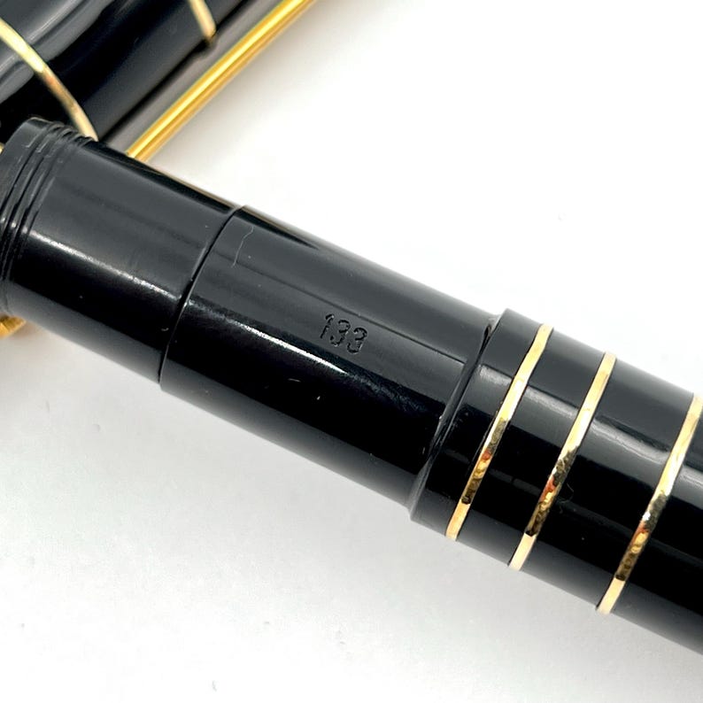 Puede incluir: Primer plano de una pluma estilogr&aacute;fica negra con detalles dorados. El bol&iacute;grafo tiene un acabado brillante y bandas doradas. El n&uacute;mero "133" es visible en el cuerpo del bol&iacute;grafo.