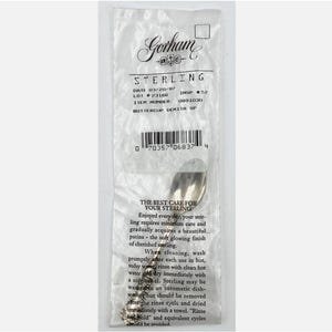 May include: A sterling silver demitasse spoon in a clear plastic Gorham package. The package has printed text including "STERLING" and "BUTTERCUP DEMITA SP". The spoon is partially visible, reflecting light.