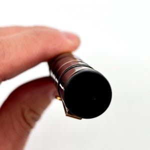 Puede incluir: Primer plano de un bol&iacute;grafo negro y dorado. El bol&iacute;grafo tiene una punta negra y un clip dorado. El cuerpo del bol&iacute;grafo es de color marr&oacute;n oscuro con detalles dorados. El bol&iacute;grafo est&aacute; en la mano de una persona.