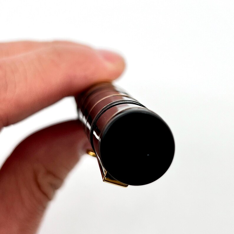 Puede incluir: Primer plano de un bol&iacute;grafo negro y dorado. El bol&iacute;grafo tiene una punta negra y un clip dorado. El cuerpo del bol&iacute;grafo es de color marr&oacute;n oscuro con detalles dorados. El bol&iacute;grafo est&aacute; en la mano de una persona.