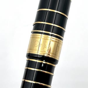 Puede incluir: Primer plano de un bol&iacute;grafo negro con detalles dorados. El bol&iacute;grafo tiene un acabado brillante y una banda dorada con la palabra "OMAN" grabada. El dise&ntilde;o del bol&iacute;grafo incluye m&uacute;ltiples bandas doradas.