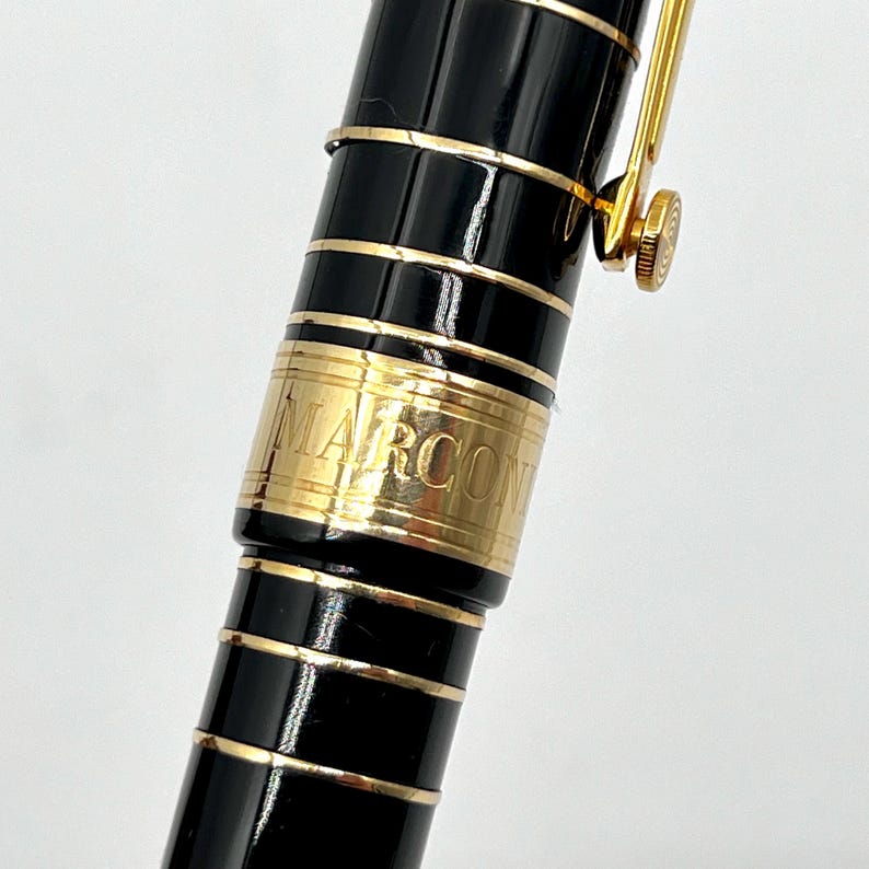 Puede incluir: Primer plano de un bol&iacute;grafo MARCON negro y dorado. El bol&iacute;grafo tiene un cuerpo negro brillante con detalles y adornos dorados. La palabra "MARCON" est&aacute; grabada en una banda dorada. El clip y el plum&iacute;n del bol&iacute;grafo tambi&eacute;n son dorados.