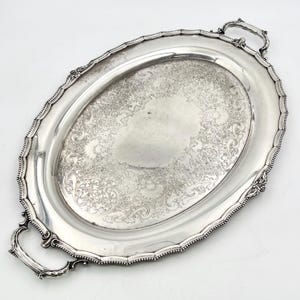 May include: An oval silver serving tray featuring intricate floral and scrollwork engravings in the center. The tray has a scalloped, beaded edge and two ornate handles on opposite ends, providing a polished metallic finish throughout the piece.
