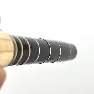 Puede incluir: Primer plano de un bol&iacute;grafo con un cuerpo negro y detalles dorados. El dise&ntilde;o del bol&iacute;grafo presenta una serie de anillos negros y dorados, creando una apariencia elegante. El bol&iacute;grafo se sostiene sobre un fondo blanco.