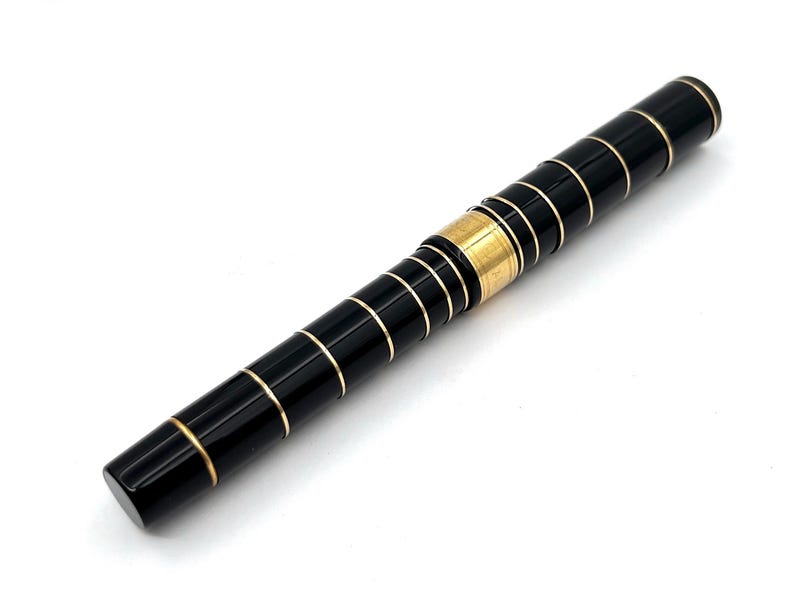 Puede incluir: Un bol&iacute;grafo negro con bandas y detalles dorados. El bol&iacute;grafo tiene forma cil&iacute;ndrica con punta redondeada. Las bandas doradas est&aacute;n espaciadas uniformemente a lo largo del cuerpo del bol&iacute;grafo, a&ntilde;adiendo un toque de elegancia. El bol&iacute;grafo est&aacute; sobre un fondo blanco.