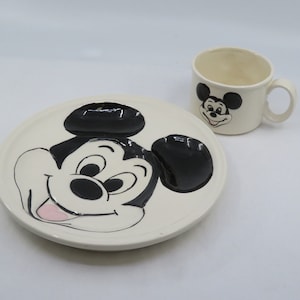 May include: A white ceramic plate and cup set featuring a black and white cartoon image of Mickey Mouse. The plate has a raised image of Mickey Mouse's face and ears. The cup has a smaller image of Mickey Mouse's face.
