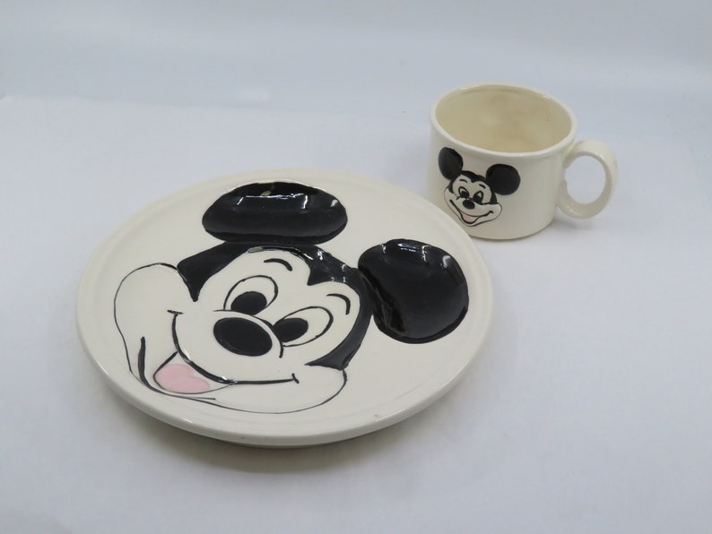 May include: A white ceramic plate and cup set featuring a black and white cartoon image of Mickey Mouse. The plate has a raised image of Mickey Mouse's face and ears. The cup has a smaller image of Mickey Mouse's face.