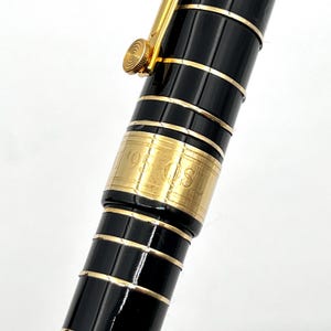 Puede incluir: Primer plano de un bol&iacute;grafo negro y dorado. El bol&iacute;grafo tiene un cuerpo negro con detalles dorados y un clip dorado. El bol&iacute;grafo tiene una banda dorada con el texto "95 OS" grabado. Es un instrumento de escritura de lujo.