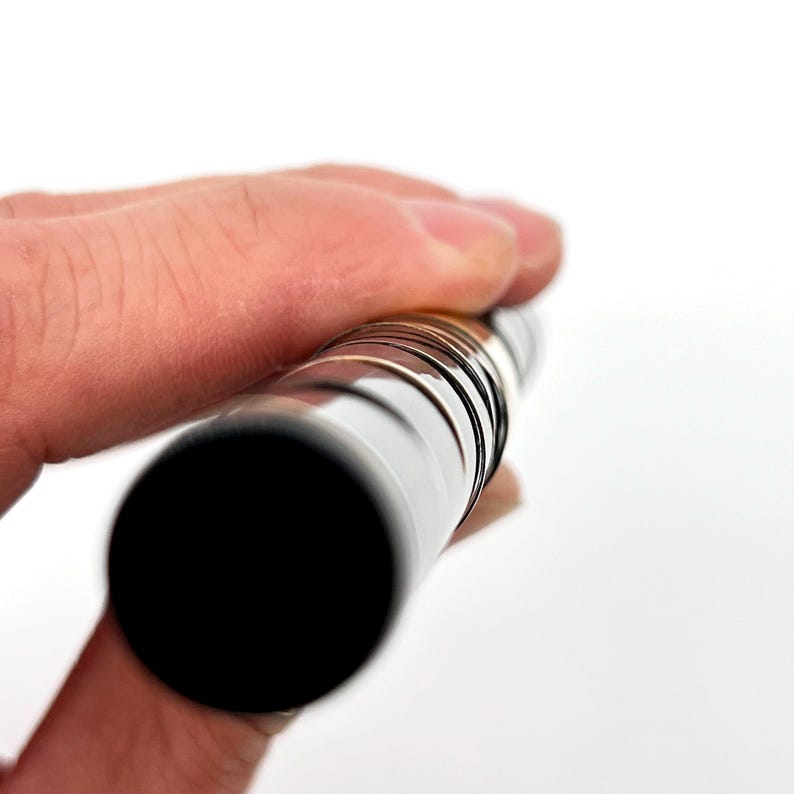 Puede incluir: Un objeto cil&iacute;ndrico con un extremo negro y un cuerpo plateado y estriado se sostiene en una mano sobre un fondo blanco. El objeto parece ser de metal y tiene una superficie reflectante. Las estr&iacute;as crean un patr&oacute;n a lo largo del cuerpo.