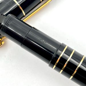 Puede incluir: Primer plano de una pluma estilogr&aacute;fica negra con detalles dorados. La pluma tiene una superficie brillante y reflectante y la palabra "ITALY" est&aacute; grabada en el cuerpo. El dise&ntilde;o de la pluma incluye bandas doradas.