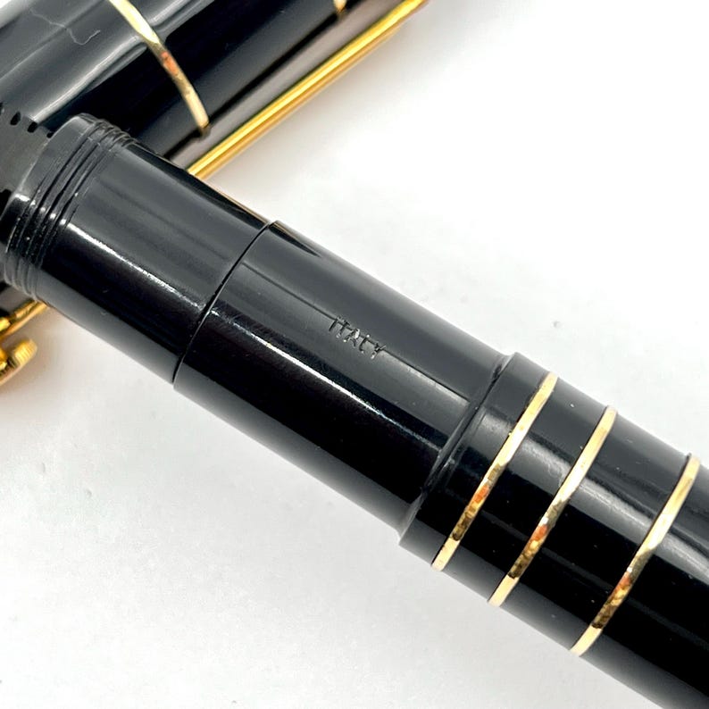 Puede incluir: Primer plano de una pluma estilogr&aacute;fica negra con detalles dorados. La pluma tiene una superficie brillante y reflectante y la palabra "ITALY" est&aacute; grabada en el cuerpo. El dise&ntilde;o de la pluma incluye bandas doradas.