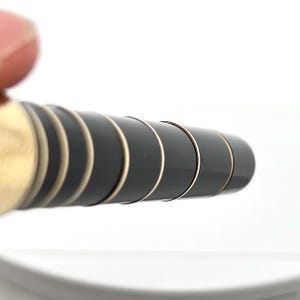 Puede incluir: Primer plano de un bol&iacute;grafo negro y dorado. El bol&iacute;grafo tiene un cuerpo negro brillante con bandas y detalles dorados. El dise&ntilde;o es elegante y moderno, con un enfoque en el contraste entre los elementos negros y dorados. El bol&iacute;grafo est&aacute; sobre un fondo blanco.