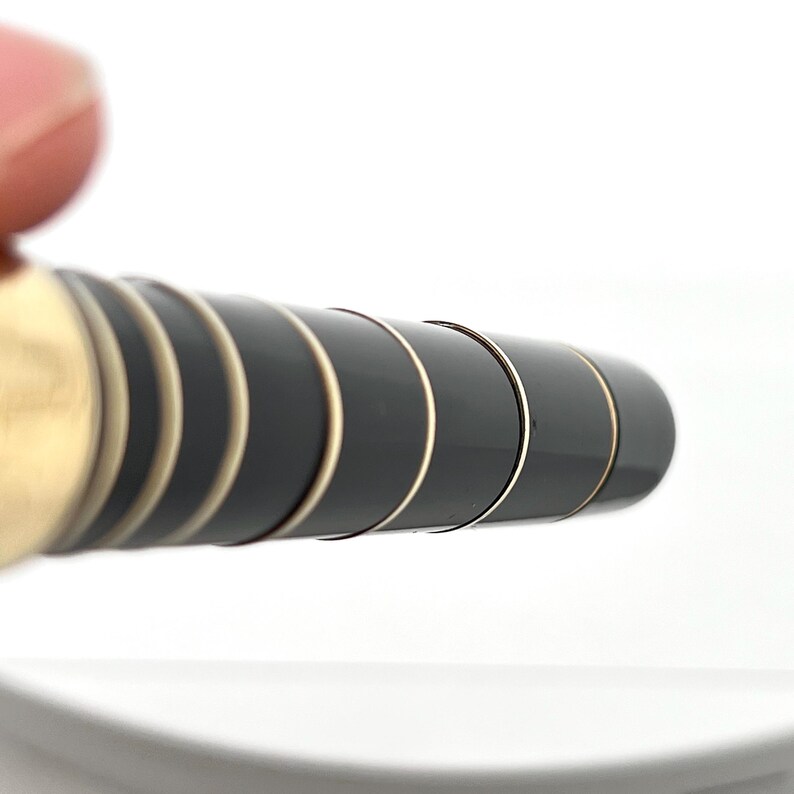 Puede incluir: Primer plano de un bol&iacute;grafo negro y dorado. El bol&iacute;grafo tiene un cuerpo negro brillante con bandas y detalles dorados. El dise&ntilde;o es elegante y moderno, con un enfoque en el contraste entre los elementos negros y dorados. El bol&iacute;grafo est&aacute; sobre un fondo blanco.