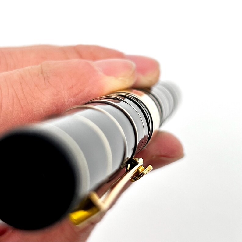 Puede incluir: Un bol&iacute;grafo negro y plateado con un clip dorado. El bol&iacute;grafo tiene una serie de anillos plateados alrededor del cuerpo. El clip est&aacute; sujeto al bol&iacute;grafo con un peque&ntilde;o tornillo. El bol&iacute;grafo se sostiene en una mano sobre un fondo blanco.