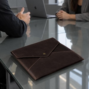 May include: A dark brown leather document folder with an envelope-style flap and a single metallic snap closure. The folder features a smooth, matte finish and stitched edges, resting on a reflective glass surface in a professional office setting.