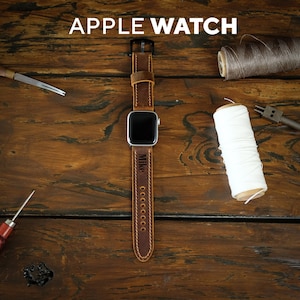 May include: A brown leather Apple Watch strap with the name "Mike" engraved. The strap is stitched with a lighter thread and has a black buckle. The text "APPLE WATCH" is at the top of the image. The watch is on a wooden surface.