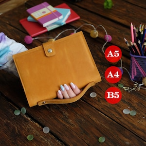 May include: A mustard yellow leather notebook cover featuring a snap button closure and a matching carrying strap. The cover is displayed alongside a purple pencil holder filled with colored pencils, small notebooks, and red circular labels marked A5, A4, and B5.