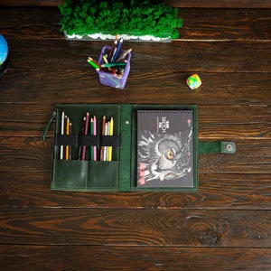 May include: A green leather art supply organizer with a sketchbook featuring an owl illustration. The organizer holds various colored pencils and pens. A small purple container with pencils and a colorful cube are also on the wooden surface.