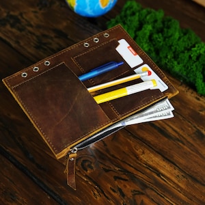 May include: A brown leather wallet with a zipper closure, containing pens, credit cards, and cash. The wallet has a distressed finish and metal eyelets along the top edge. The background includes a wooden surface, a globe, and green moss.
