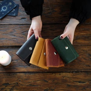 May include: A collection of four small leather wallets in black, mustard yellow, reddish-brown, and dark green. Each wallet features a simple flap design secured with a single silver-toned metal snap button closure, showcasing a smooth matte finish.