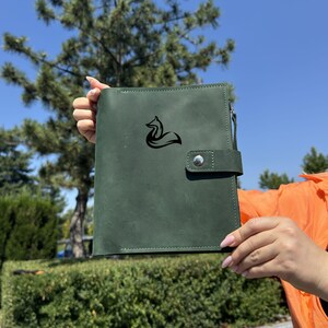 May include: A dark green leather journal with a black fox logo. The journal has a zipper closure and a snap closure. The background is a blue sky and green foliage.