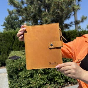 May include: A tan leather journal with a snap closure and a strap. The word "Emily" is engraved on the lower right corner. The journal is held up against a background of green foliage and a blue sky.