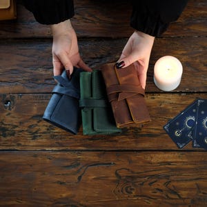 May include: Three leather tarot card holders in navy, green, and brown, each secured with a strap. A lit white candle and two tarot cards with a celestial design are also visible on a wooden surface.