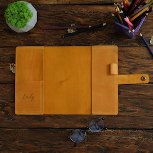 May include: A yellow leather folder with a flap closure and a personalised name, "Emily", engraved on the front. The folder is open and lying on a wooden surface.
