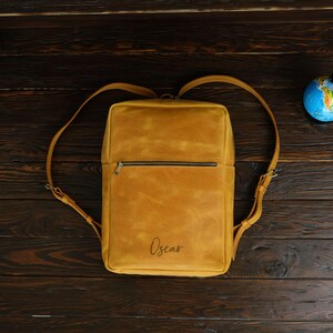 May include: A tan leather backpack with a zippered pocket and the name "Oscar" embossed on the front.