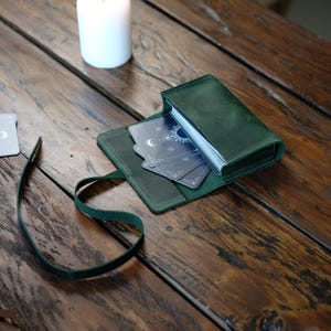 Pode incluir: Uma bolsa de couro verde-escura contendo um baralho de tarot com desenhos celestiais de sol, lua e estrelas. A bolsa possui um fecho de tira de couro envolvente e est&aacute; parcialmente aberta, revelando o baralho de cartas no seu interior.