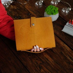 May include: A brown leather portfolio with a silver button closure. The portfolio is being held by a hand with white nail polish.
