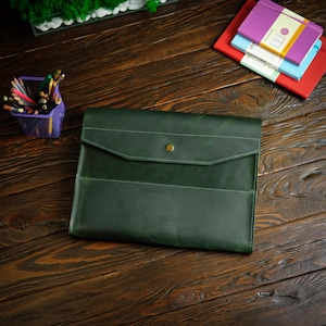 May include: A dark green leather document folder featuring a flap closure with a single brass-colored snap button, visible white stitching along the edges, and a smooth matte finish. The folder is rectangular with a structured design.