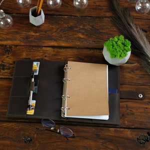May include: A brown leather bound journal with a ring binder and a blank page open. The journal is on a wooden table with a small potted plant, a pen, and glasses.