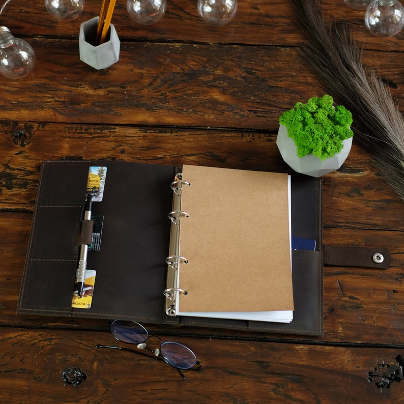 May include: A brown leather bound journal with a ring binder and a blank page open. The journal is on a wooden table with a small potted plant, a pen, and glasses.