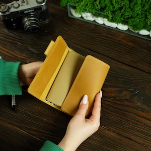 May include: A mustard-coloured leather journal with a matching pen. The journal is open, revealing a tan-coloured notebook inside. The journal is held by a person with a green sleeve. A vintage camera and decorative moss are in the background.