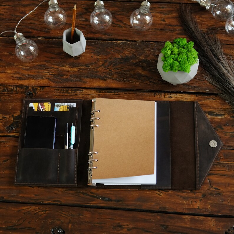 May include: A brown leather bound journal with a ring binder, a pen, and credit cards in the pockets. The journal is open to a blank page with a brown paper insert.