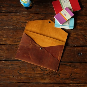 May include: A brown leather envelope with a flap that folds over. The envelope is open and has a yellow leather interior. The envelope is on a wooden surface.