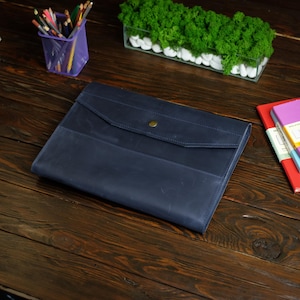 May include: A dark blue leather document folder featuring a flap closure secured with a single brass-colored metal snap button. The rectangular case has visible stitching along the edges and a smooth, matte finish on the front surface.
