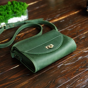 May include: A dark green leather crossbody bag with a flap closure and a brass buckle. The bag has a long strap and is sitting on a wooden surface.