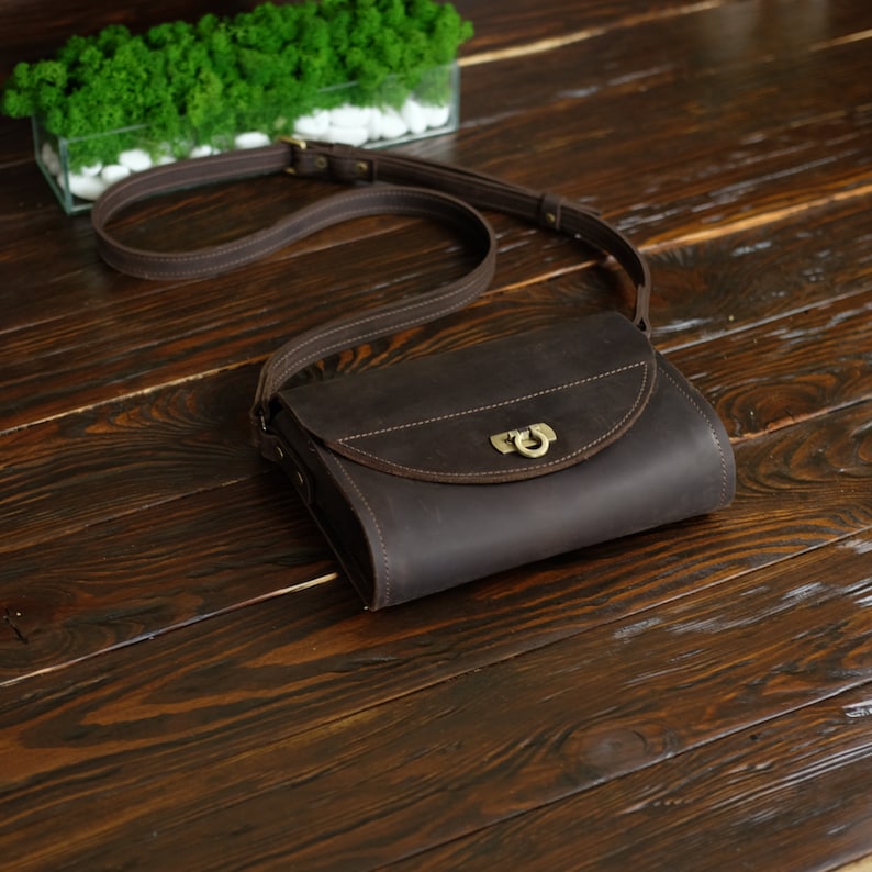 May include: A brown leather crossbody bag with a flap closure and a brass buckle. The bag has a long strap and is sitting on a wooden surface.