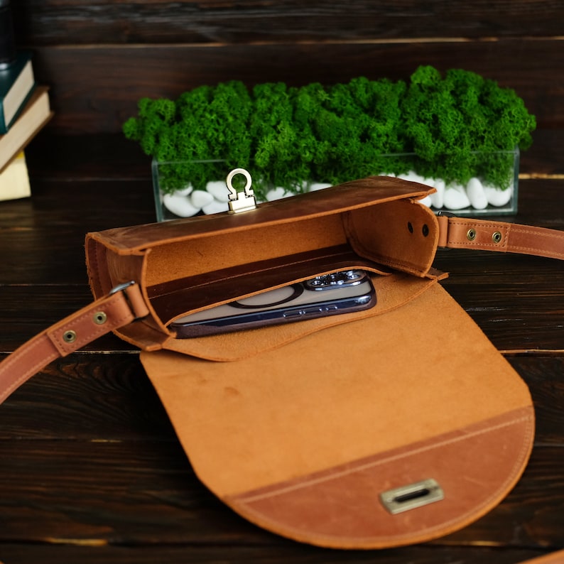 May include: A brown leather satchel bag with a flap closure and a gold metal buckle. The bag is open and a smartphone is inside.