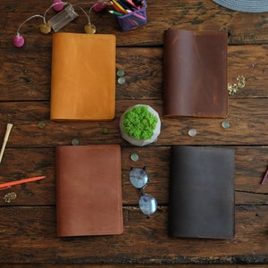 Puede incluir: Cuatro cuadernos encuadernados en cuero de diferentes colores: naranja, marr&oacute;n, marr&oacute;n oscuro y negro. Los cuadernos est&aacute;n dispuestos sobre una superficie de madera con una peque&ntilde;a planta en maceta en el centro.