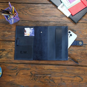 May include: A dark blue leather organizer folio featuring multiple interior pockets, a pen loop, and a zippered compartment. The left side displays the embossed initials E.W. and holds two small cards, while a white smartphone rests on the right side.
