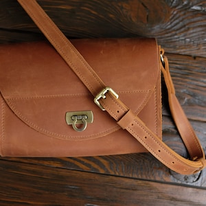 May include: A brown leather satchel bag with a flap closure and a brass buckle. The bag has a long adjustable strap.